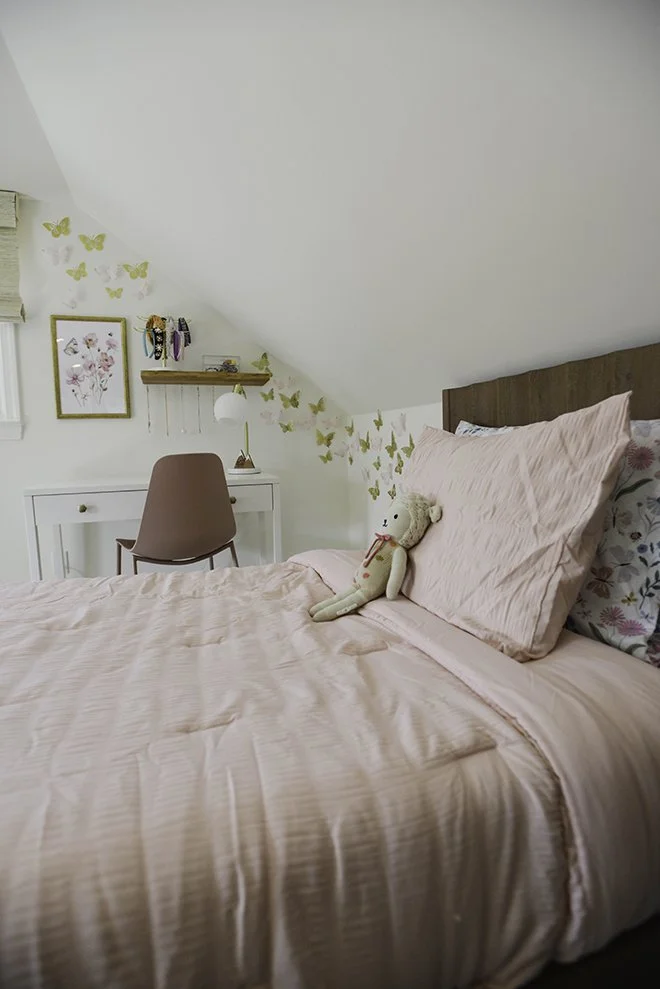A bedroom with a pink bedspread, matching pillows, and a teddy bear on the bed. There is a white desk with a pink chair, a framed floral artwork, a shelf with decorations, and butterfly wall decals near the sloped ceiling.