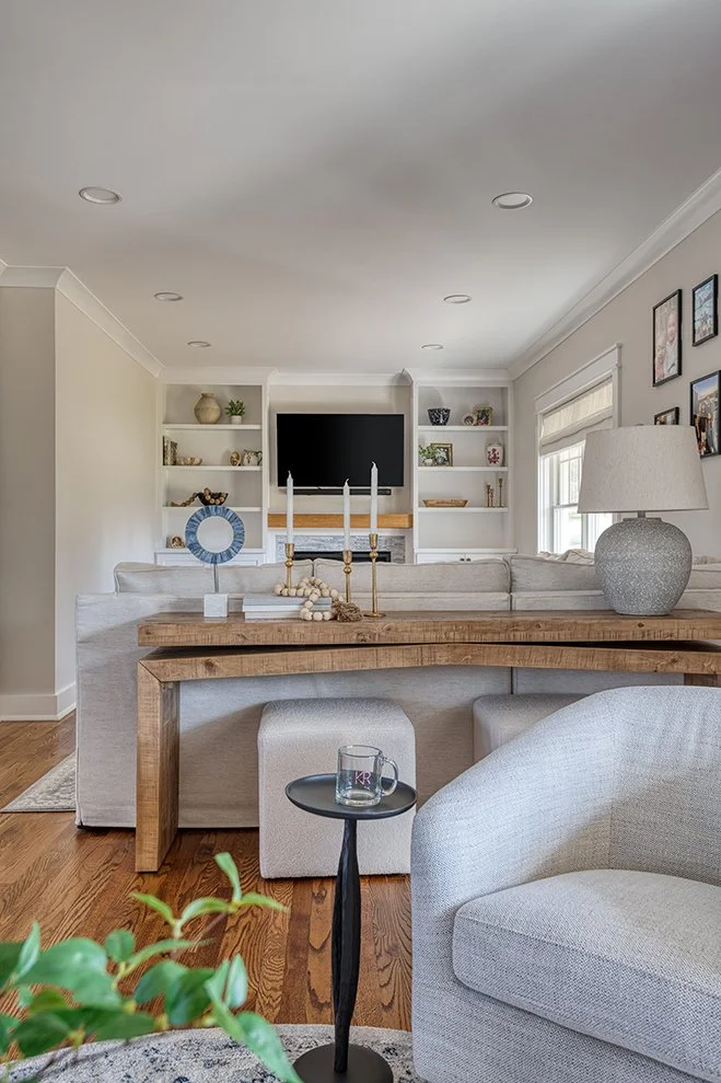Living room with white walls, built-in shelves, a flat-screen TV, beige sofa, wooden coffee table, gray armchair, small black side table with a glass mug, large gray table lamp, and framed pictures on the wall.