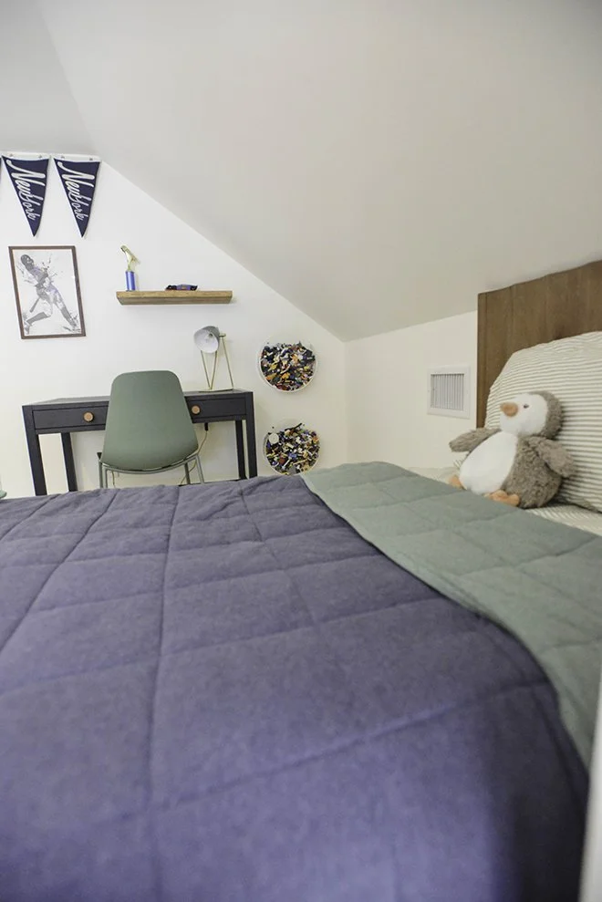 A bedroom with a bed that has a dark blue and green quilt, a stuffed penguin toy on the pillow, a desk with a green chair, two decorative Lego brick circular wall hangings, a small desk lamp, and two blue pennants hanging on the wall.