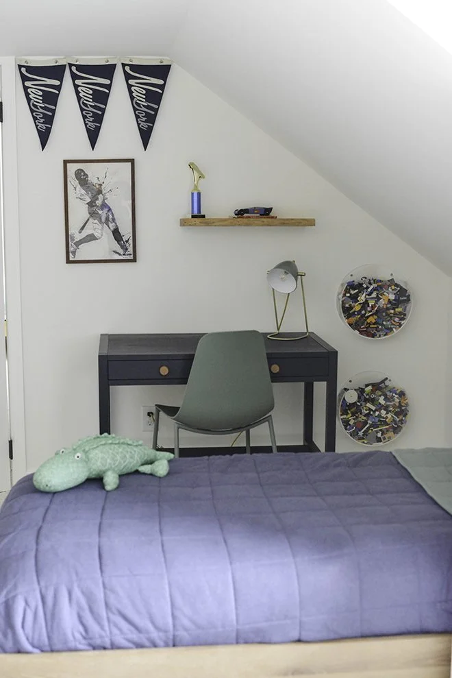 A bedroom with a bed covered in a blue quilt, a green plush crocodile toy on it, a dark desk with a gray chair, a picture of a hockey player, a wooden shelf with a trophy and small toy, a stylish desk lamp, and two circular wall-mounted LEGO brick co