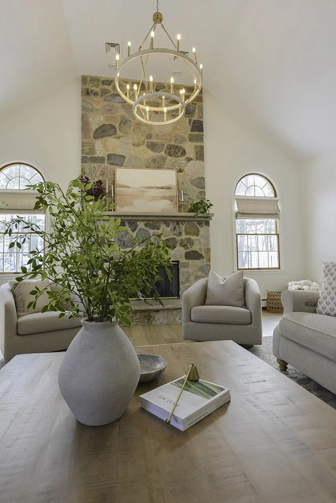 Living room with a stone fireplace, two arched windows, beige armchairs, a light wood coffee table with a vase of green leaves, a book, and a modern chandelier.