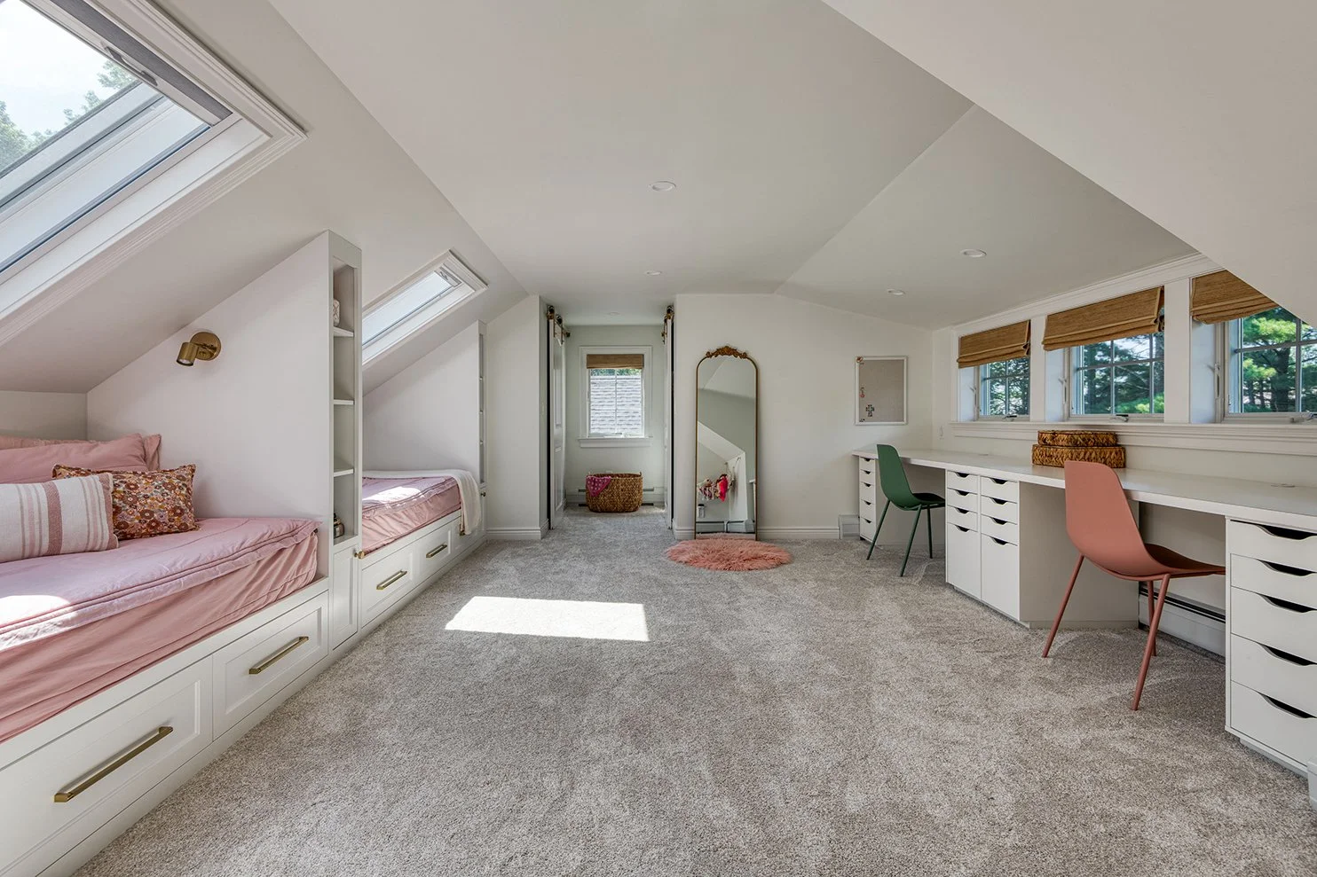A spacious attic bedroom with white walls and ceiling, beige carpeting, built-in pink beds with pillows on the left, a full-length mirror and small window with blinds in the center, and a long white desk with pink and green chairs on the right.
