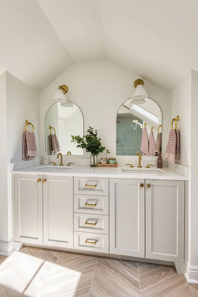 White bathroom vanity with two sinks and gold fixtures, including faucets and towel rings, set against a white wall with two arched mirrors and pink checkered towels. A plant and small decorative items are on the countertop, with beige and brown patt