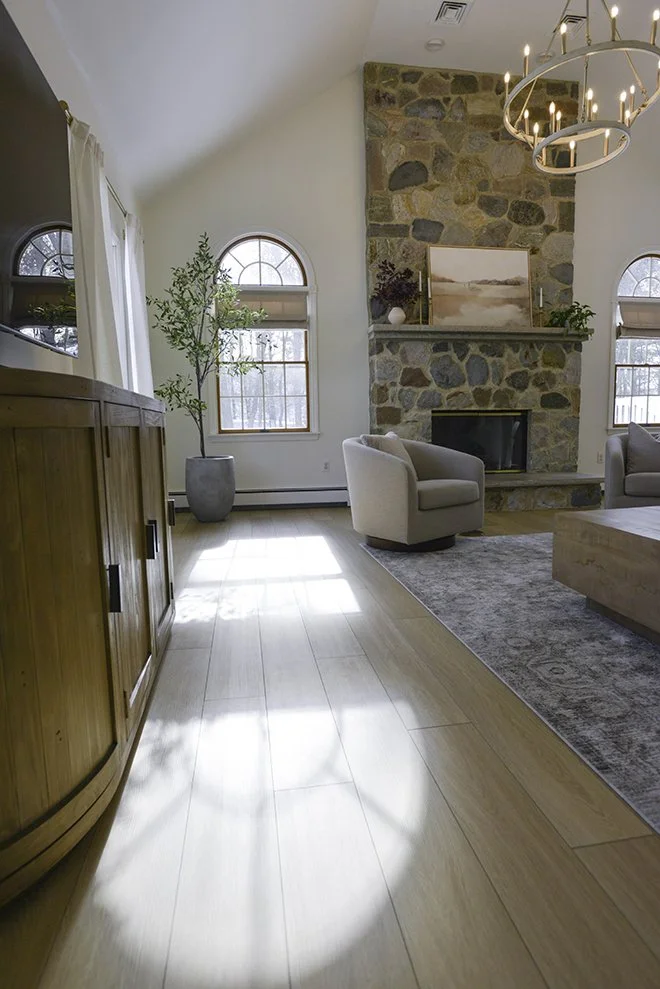 Living room with a stone fireplace, two arched windows, a beige armchair, potted plant, chandelier, wooden furniture, and a gray rug.