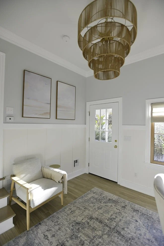 A cozy living room corner with a white armchair, light-colored throw blanket, a small side table, a patterned rug, a white door with glass window, a window with blinds, light gray walls, framed artwork, hardwood floors, and a multi-layered hanging ch