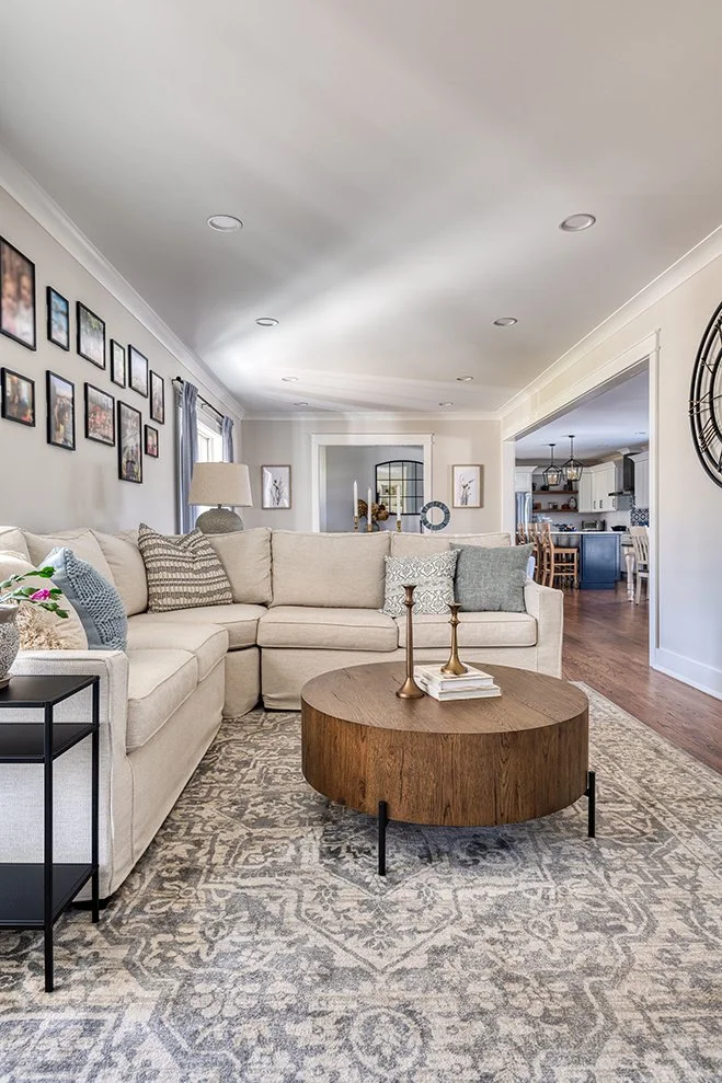 Living room with beige sectional sofa, patterned throw pillows, round wooden coffee table, gray and white area rug, and photo frames on the wall.