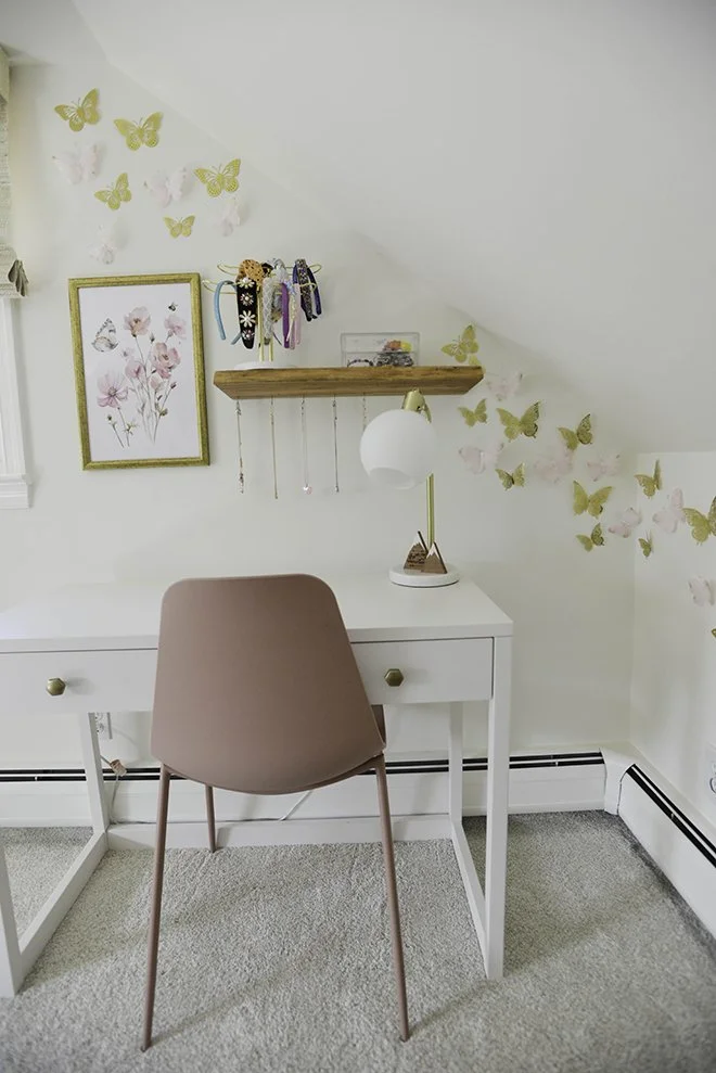 A white desk with a pink chair, a white and gold desk lamp, a wooden shelf with jewelry, wall art of pink flowers and yellow butterflies, and a beige carpet in a room with a white wall.