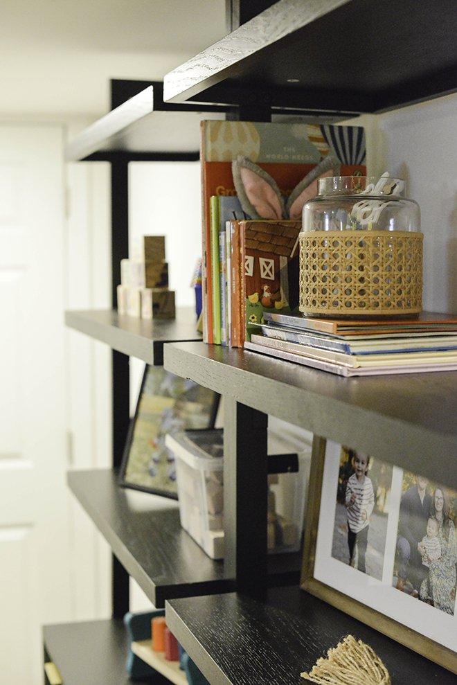 Close-up of a black wooden shelf with colorful books, a glass jar with a woven holder, a framed family photo, and decorative items.