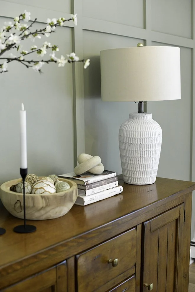 Decorative wooden sideboard with a white ceramic table lamp, a tall white candle in a black holder, a bowl of decorative balls, and a small stack of books.