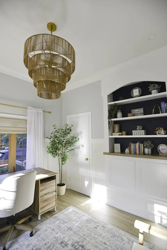 Bright living room with a gold chandelier, a white and wooden standing desk, white curtains, a potted plant, a built-in white and navy bookshelf with decorative items and books, partial view of a white chair, and a light-colored area rug.