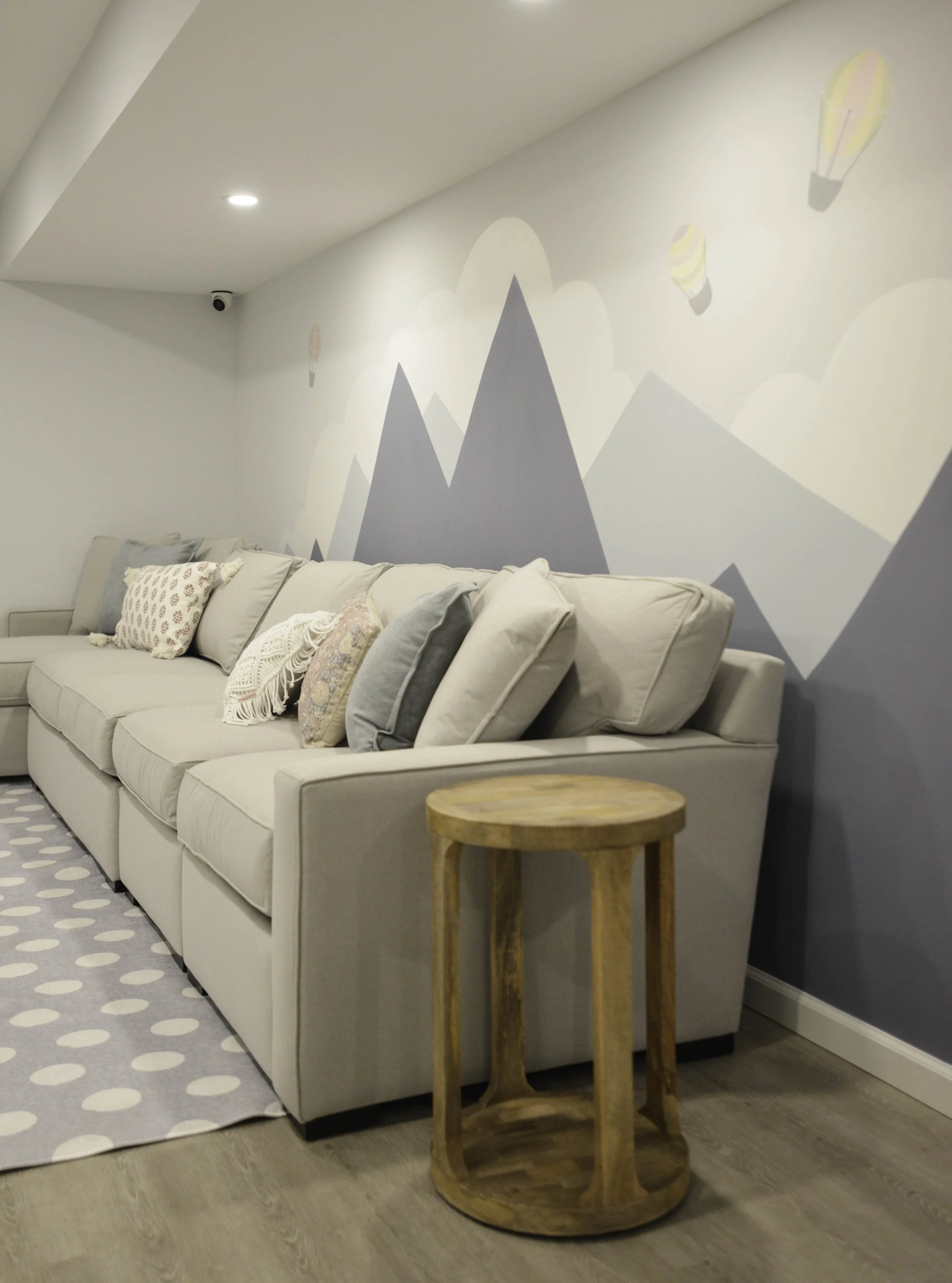 A cozy living room corner with a light gray sectional sofa adorned with decorative pillows, a small wooden side table, a polka dot rug, and a pastel mountain mural with hot air balloons painted on the wall behind.