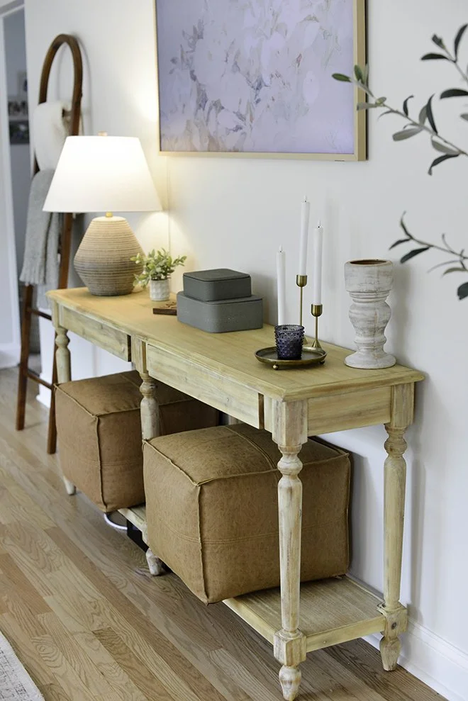 A wooden console table with two brown ottomans underneath, decorated with a table lamp, potted plant, gray storage boxes, candles, a cup, and a vase, against a white wall with a large abstract painting.