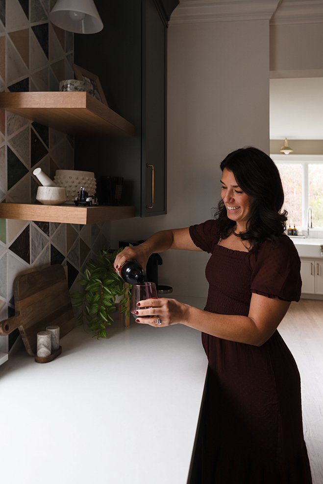 A woman in a brown dress pouring red wine into a glass in her kitchen.