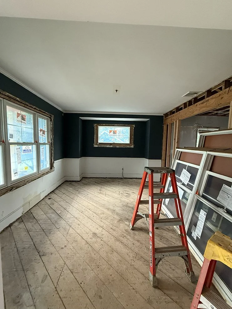 Room under renovation with new windows leaning against the wall, a red step ladder in the middle, and floorboards torn up for remodeling.