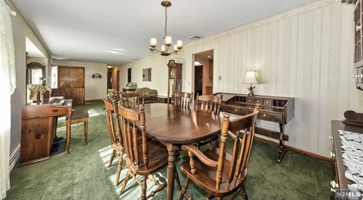 A dining room with a large wooden table and chairs, green carpet, a chandelier, and vintage furniture.