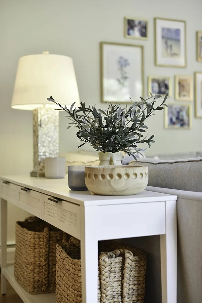 A white side table decorated with a large potted plant, two small pots, and a lamp, with woven baskets on the lower shelf and a gallery wall of framed pictures in the background.
