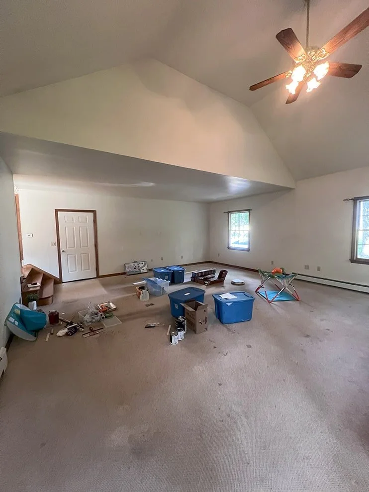 Empty living room with beige carpet, scattered storage boxes, toys, and miscellaneous items, two windows, white walls, ceiling fan with lights
