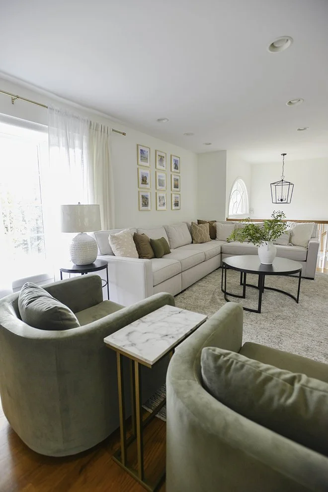 A cozy living room with light-colored sofas, a coffee table with a white vase of greenery, green armchairs, and wall art, illuminated by natural light from sliding glass doors.