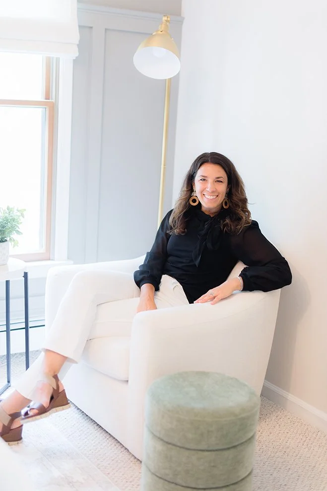 A woman sitting in a white armchair in a bright room, smiling, wearing a black top, white pants, and tan sandals, with a floor lamp and a window with a white shade behind her.
