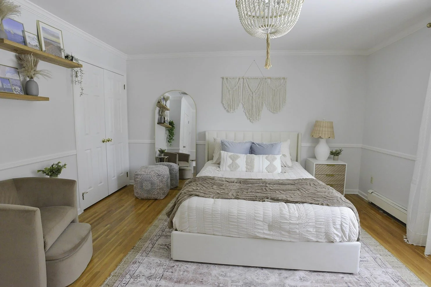 A bedroom with white walls and wooden floors. It features a white bed with a beige blanket and decorative pillows, a white nightstand with a lamp and plant, a mirror, a beige armchair, and shelves with framed pictures and decor. There's a macramé wal