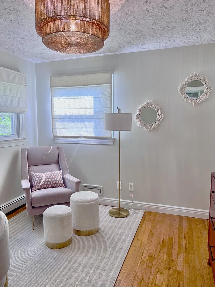A cozy corner with a pink armchair, two white ottomans, a tall floor lamp, and two decorative round mirrors on the wall. The room has light-colored walls, a window with a Roman shade, and a chandelier hanging from the ceiling. Wooden flooring is part