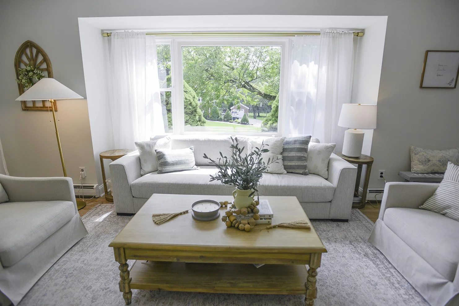 Bright living room with large window, white sofas, side tables with lamps, a wooden coffee table with a plant, candles, books, and decorative items, and views of greenery outside.