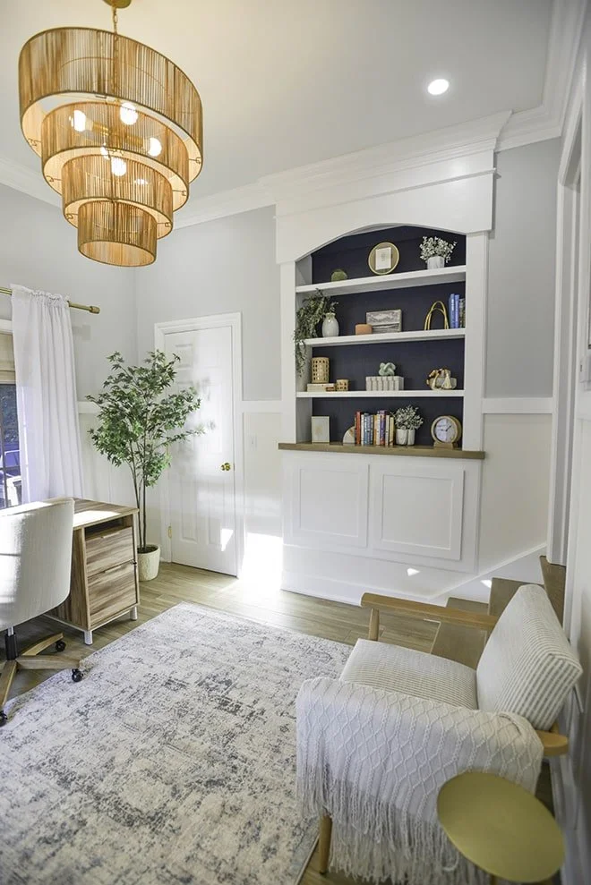 Bright living room corner with white walls, wooden flooring, a beige upholstered armchair with a throw, a wooden desk with a rolling chair, a potted plant, built-in white shelves with decorative items and books, a window with curtains, and a large ba