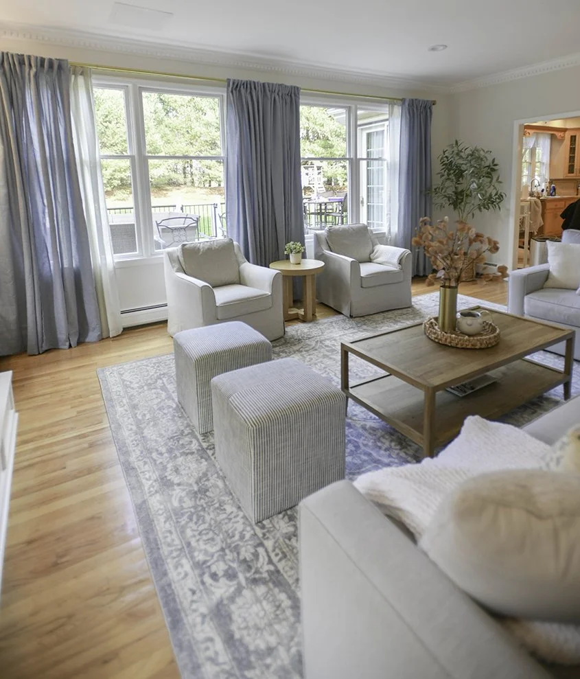 Bright living room with large windows, white armchairs, and wooden coffee table decorated with flowers, beige walls, hardwood flooring, and blue curtains.