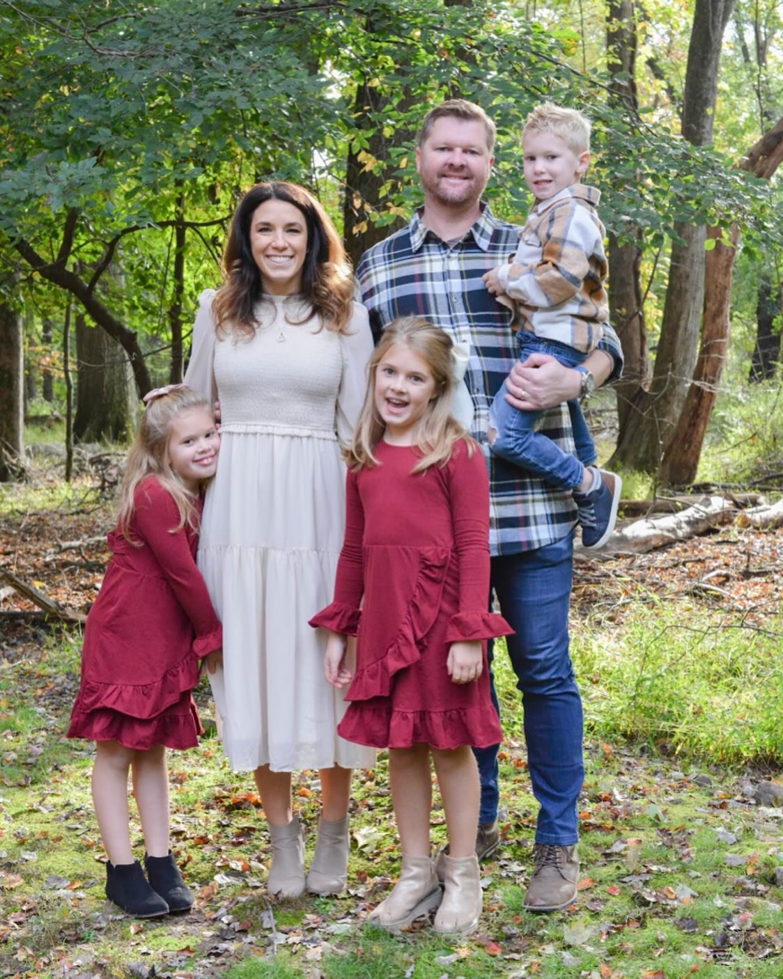Family of five in a wooded park, posing for a photo. Two young girls in red dresses and boots stand in front, a woman in a white dress and two men, one holding a young boy, stand behind them.