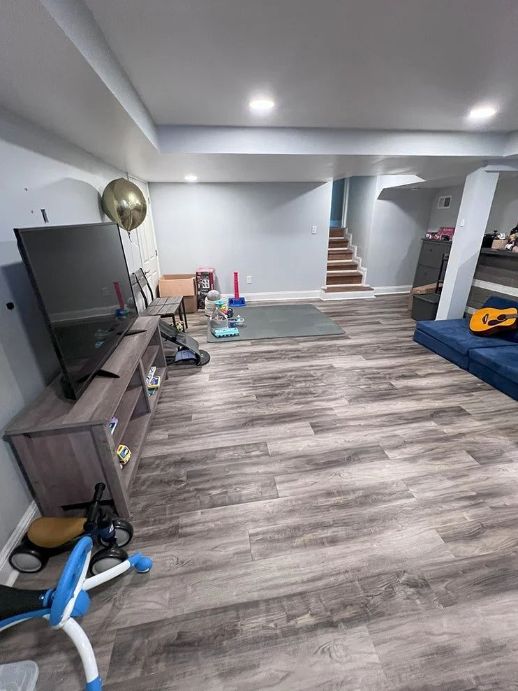 A living room with wooden flooring, a large flat-screen TV on a wooden stand, a gold balloon, a blue couch with a guitar, a small bike, and children's toys near a staircase.