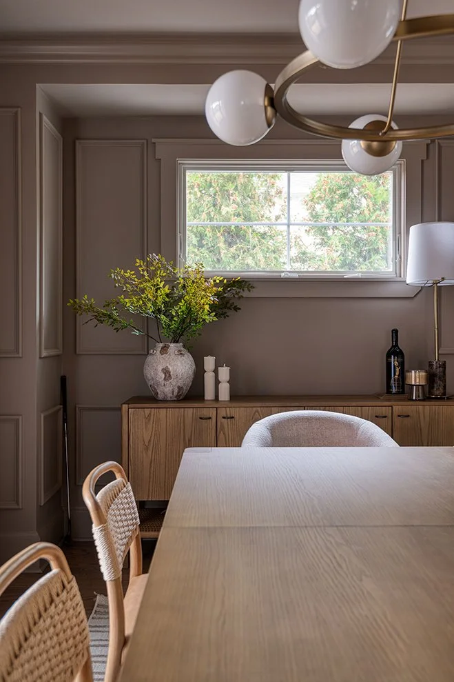 Dining room with a wooden table, wicker chairs, a sideboard with a vase of green foliage, and modern decor including a black bottle, candles, and a floor lamp. A window shows green trees outside.