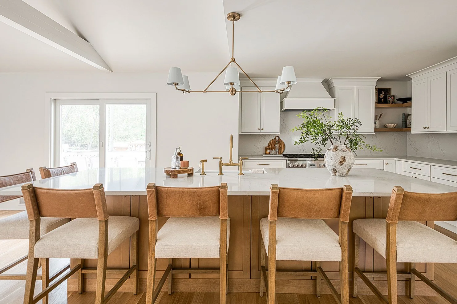 A modern kitchen with white cabinets, a white marble island, wooden barstools, and a large window door leading outside. Decor includes a large vase with greenery, a chandelier, and minimal accessories.