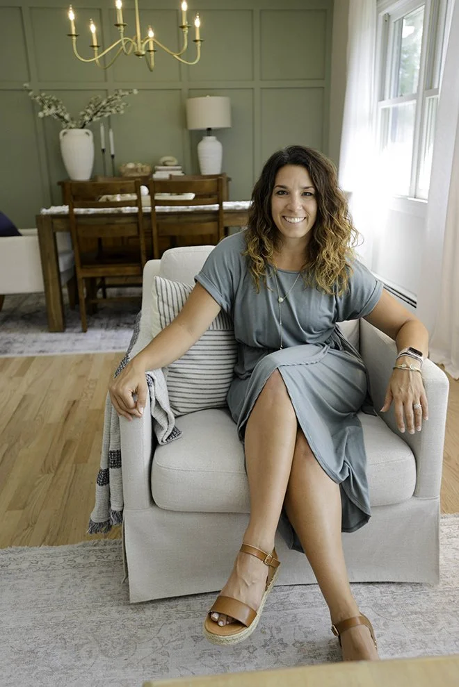 A woman with wavy brown hair sitting on a light-colored armchair in a well-lit living room, smiling at the camera. She is wearing a teal dress, sandals, and accessories. Behind her is a dining area with a wooden table, chairs, vases, and a chandelier