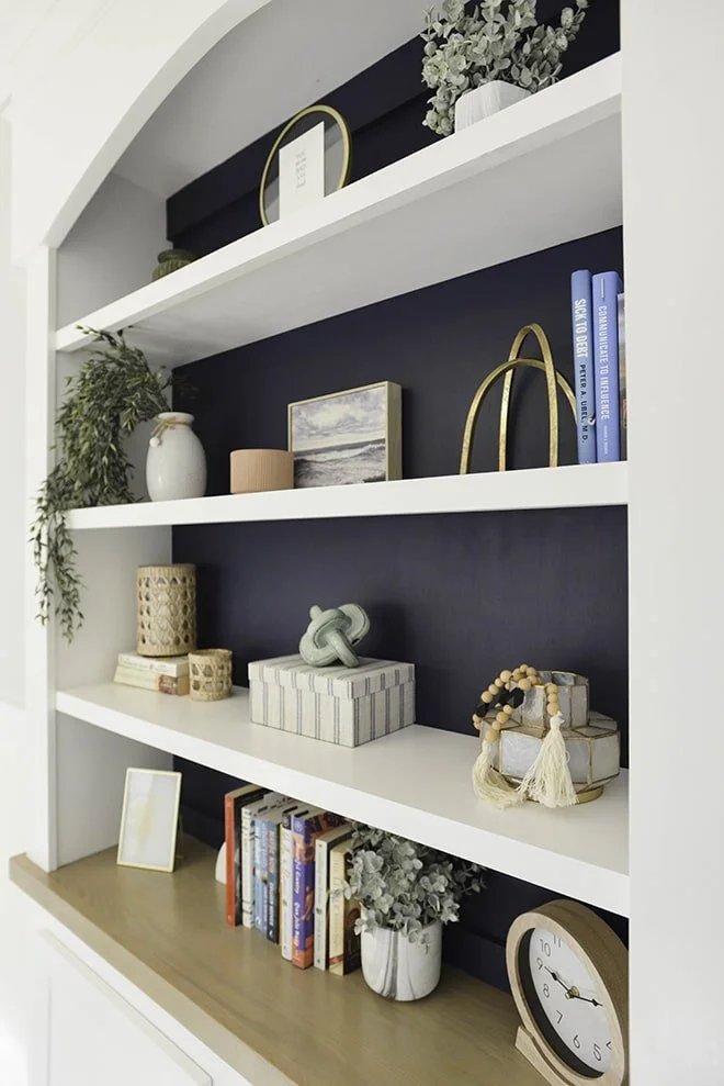 Decorative white and dark navy built-in bookshelf with plants, framed art, books, and decorative objects inside a home.