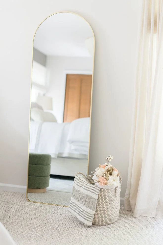 A tall, oval mirror with a thin gold frame leaning against a white wall in a bright, airy bedroom. In front of the mirror is a woven basket filled with pink and white flowers, with a striped cloth draped over the side. A window with sheer curtains is