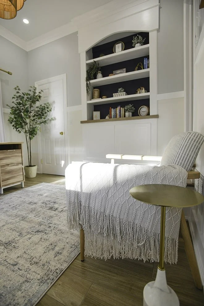 Living room with a white armchair draped with a white woven throw, a side table with a gold top and white base, a tall potted plant, a white door, a built-in bookshelf with decorative objects and books, a light-colored wooden side table, a patterned 