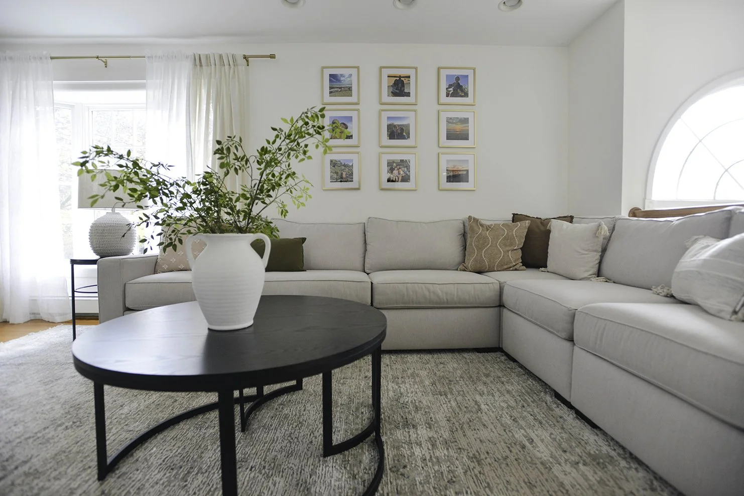 Bright living room with white sectional sofa, decorative pillows, framed photos on the wall, a large window with sheer curtains, a black coffee table with a vase of greenery, a side table with a lamp, and an arched window.