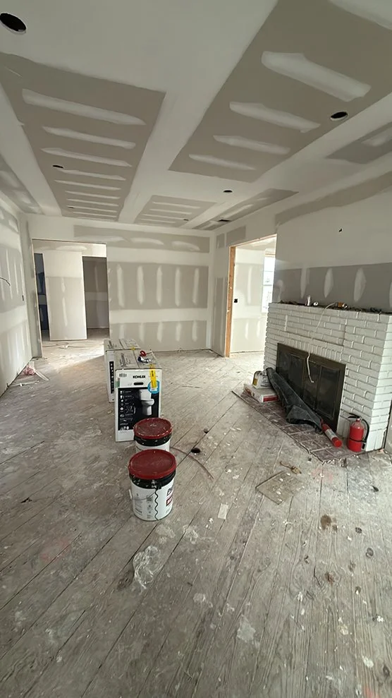 Interior of a room under construction with drywall ceilings, a brick fireplace, and construction supplies on the floor, including buckets, a box, and a fire extinguisher.