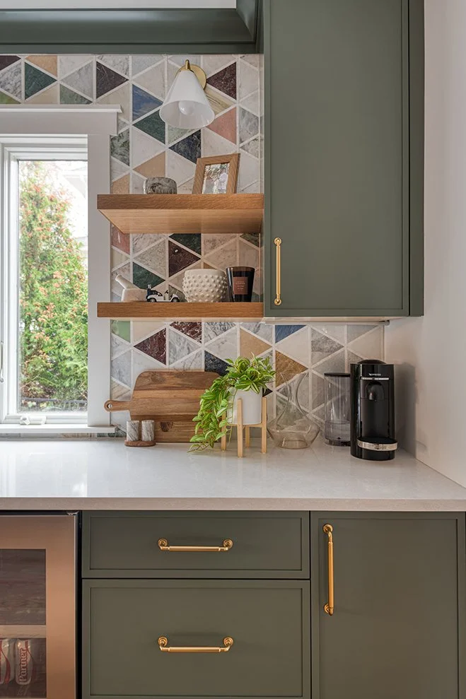 Kitchen counter with green cabinets and open wooden shelves, decorated with various dishes and small decorative items. A window shows greenery outside, and a coffee maker is on the right side of the counter. The backsplash features colorful geometric