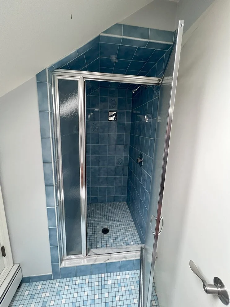 Small blue-tiled shower stall with glass door and silver frame, located in a bathroom with tiled floor and white walls.