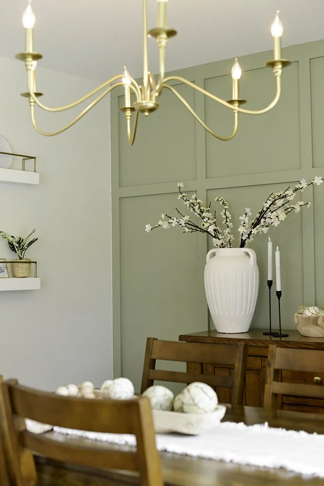 Interior dining room with a chandelier, wooden table and chairs, a large white vase with branches of white flowers, and green paneled walls.