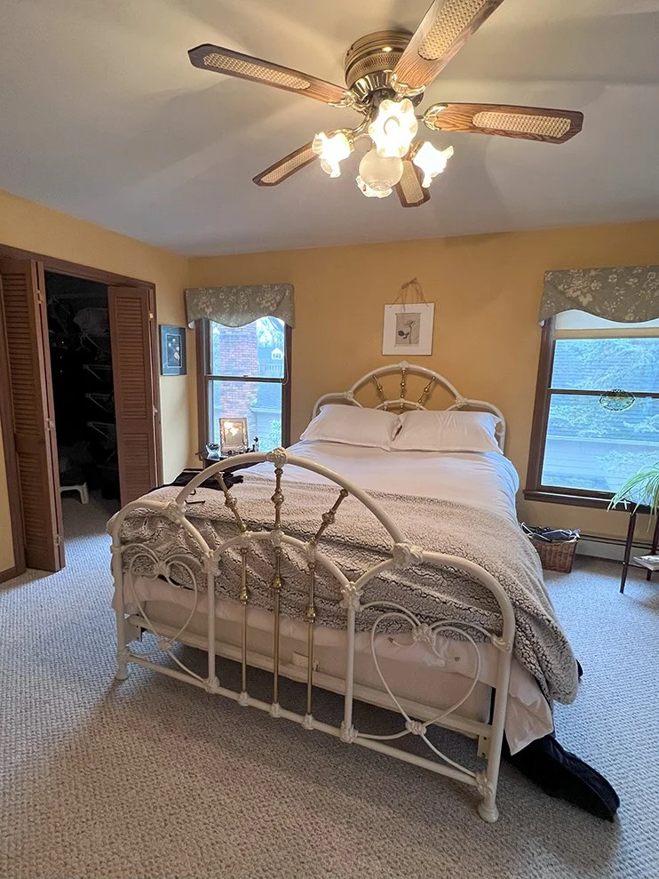 A cozy bedroom with a vintage white metal bed, beige walls, two windows with floral valances, and a ceiling fan with light fixtures. There is a small table with picture frames and a small plant near the window, and a closet with wooden louvered doors