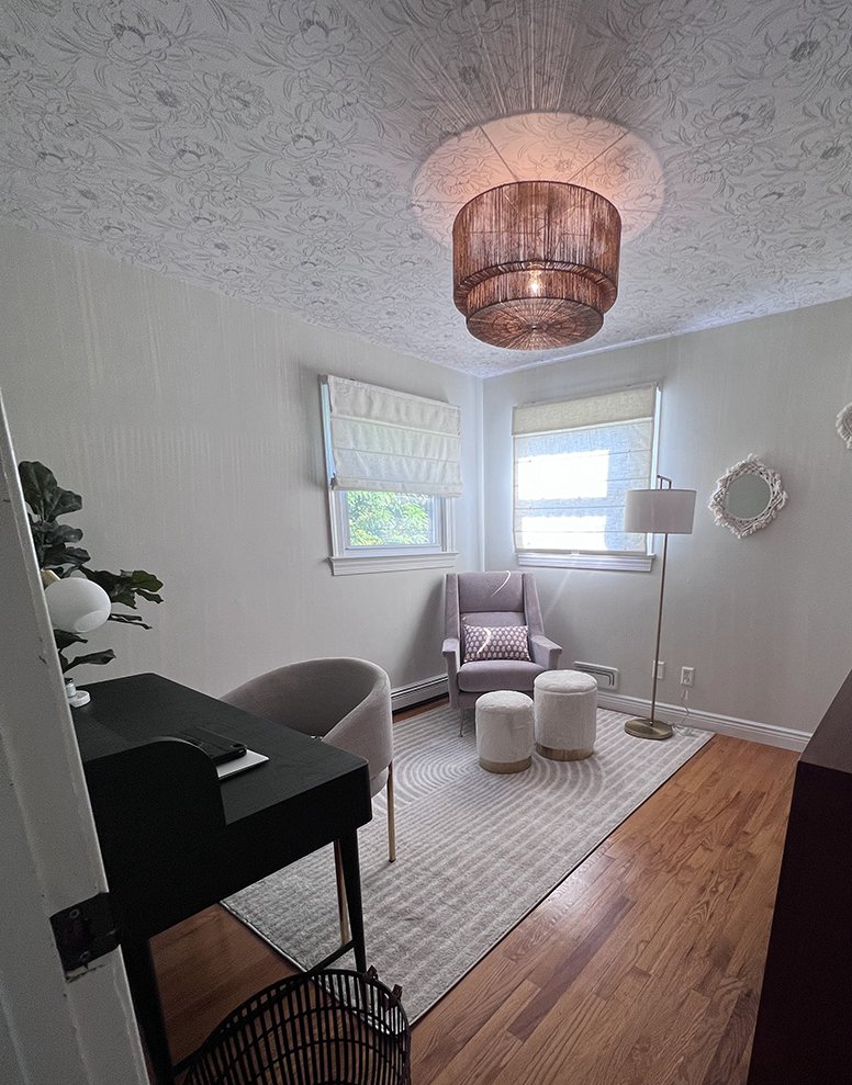 A cozy, stylish room with a gray armchair, two small matching ottomans, a floor lamp, and a decorative mirror on the wall, with natural light coming through two windows.