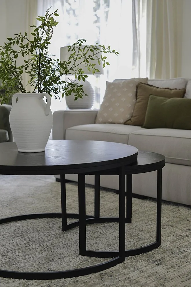 Living room with white sofa, beige and green cushions, black nested coffee tables, white vase with green plant, beige curtains, natural light from window.