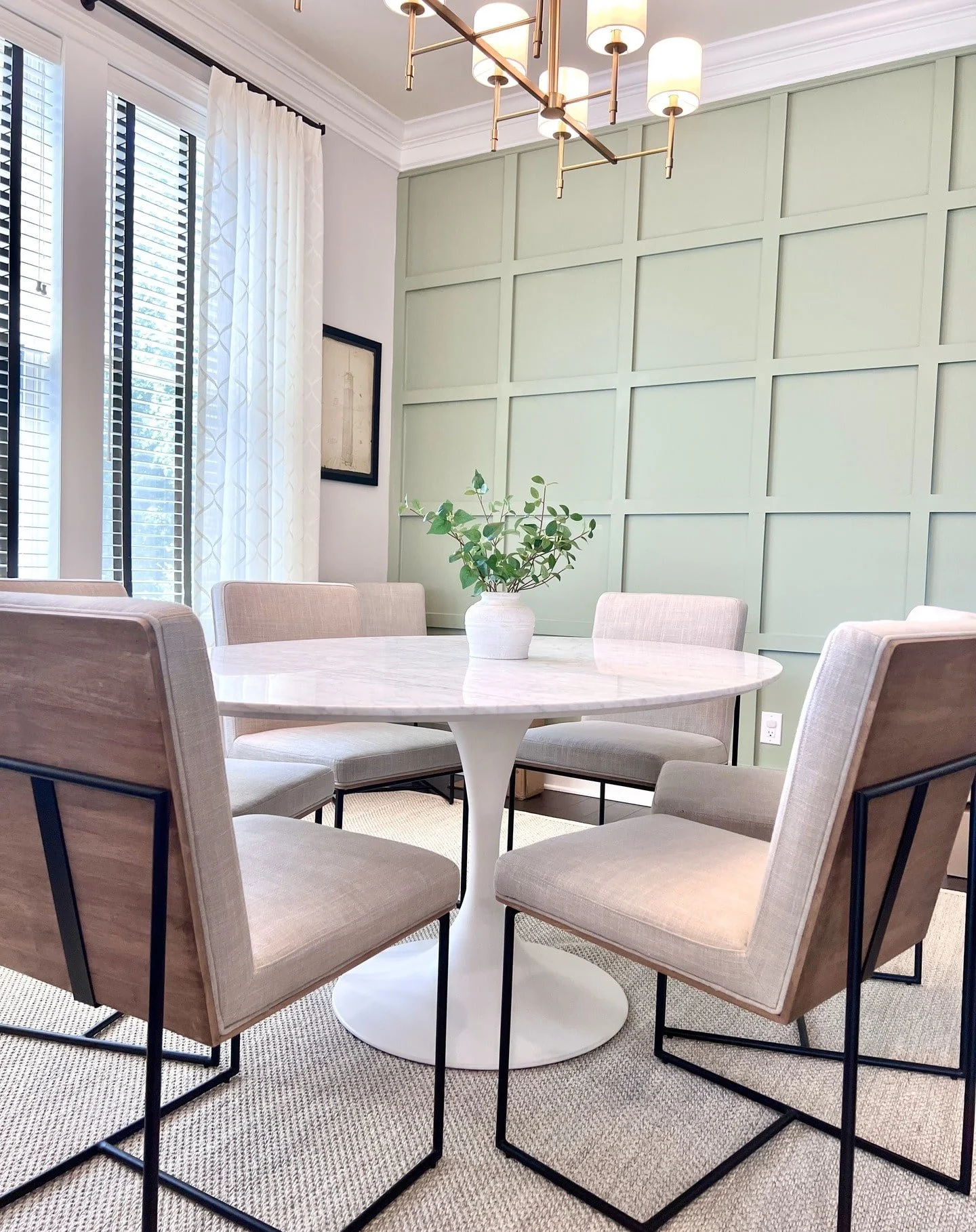 A dining room with a round white marble table, six beige upholstered chairs with black metal frames, a green paneled accent wall, white curtains, and a modern gold chandelier.