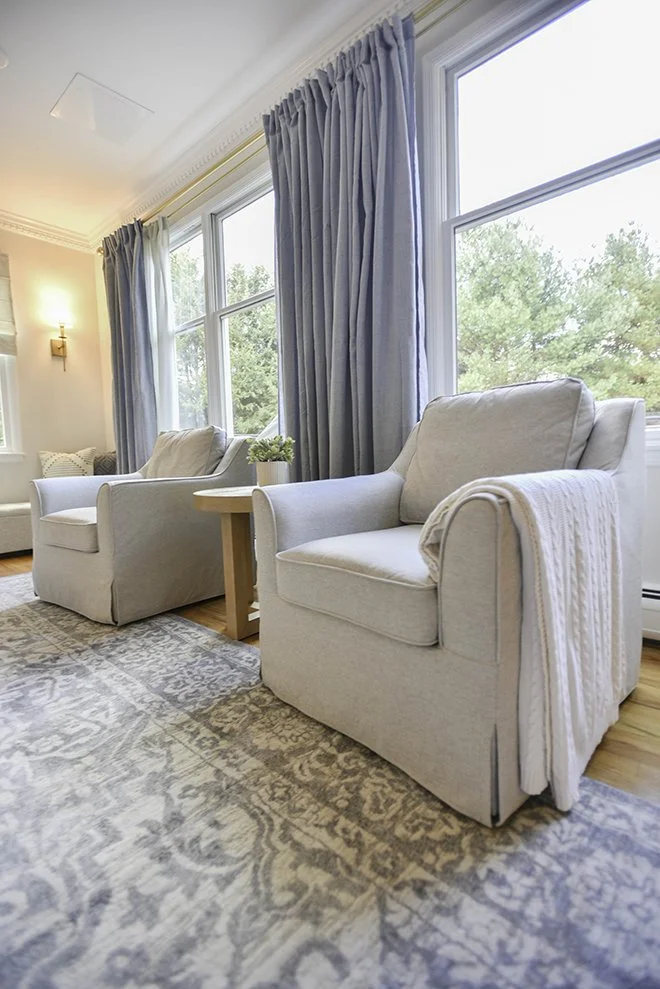 A cozy living room with two white armchairs near large windows with blue curtains, a small wooden side table with a potted plant, a patterned rug, and a glimpse of another sofa in the background.