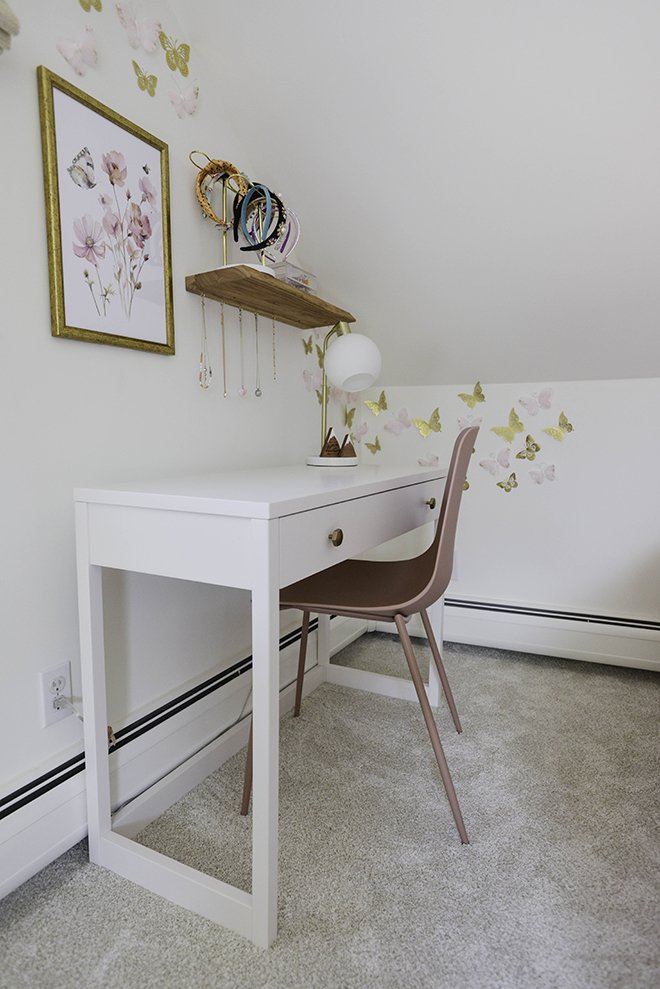 A small white desk with a drawer in a room with butterfly and flower wall decor, a pink chair, and a bookshelf with jewelry hanging on it above the desk.