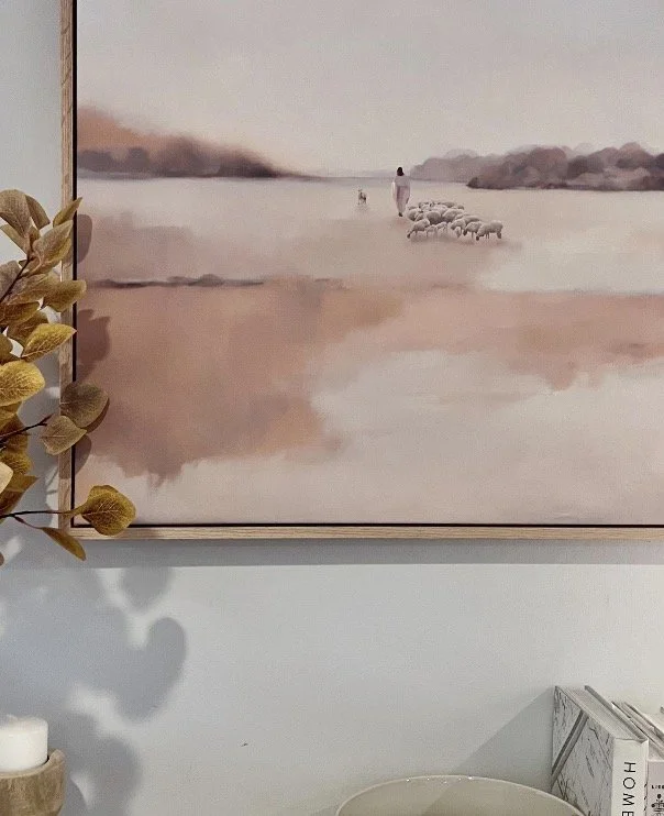 A watercolor painting of a person herding sheep across a body of water with hills in the background, hung on a wall.