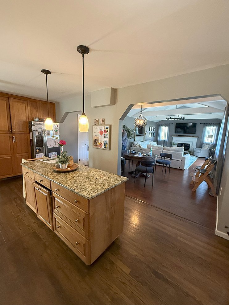 View of a kitchen with wooden cabinets and granite countertop island, two hanging pendant lights, and a doorway leading to a living room with white couches, a fireplace, and a flat-screen TV.