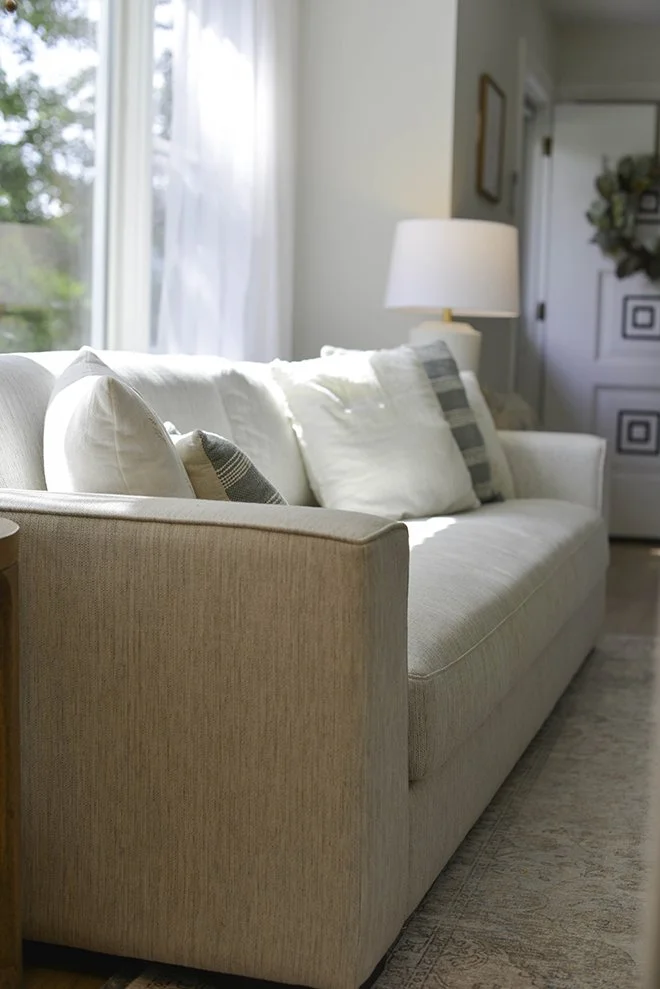 A cream-colored sofa with decorative pillows beside a window with sheer curtains, in a bright living room with a lamp and a door in the background.